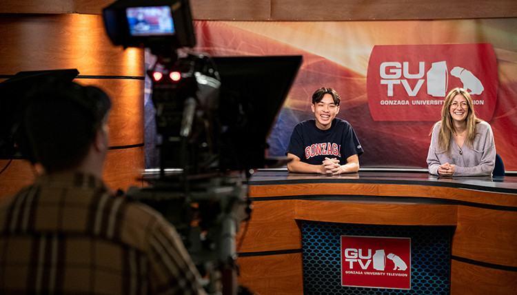 Two student broadcasters at the broadcast desk with a camera operator in the background. Two student broadcasters at the broadcast desk with a camera operator in the background.