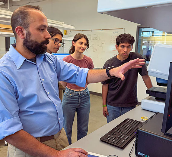 Professor gestures to a computer screen while three students stand behind him. [Ferro]