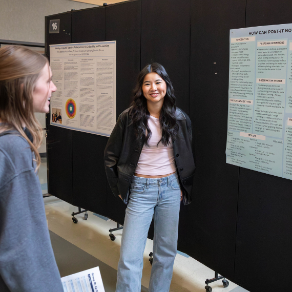 Woman stands next to poster board and smiles as she presents to another student