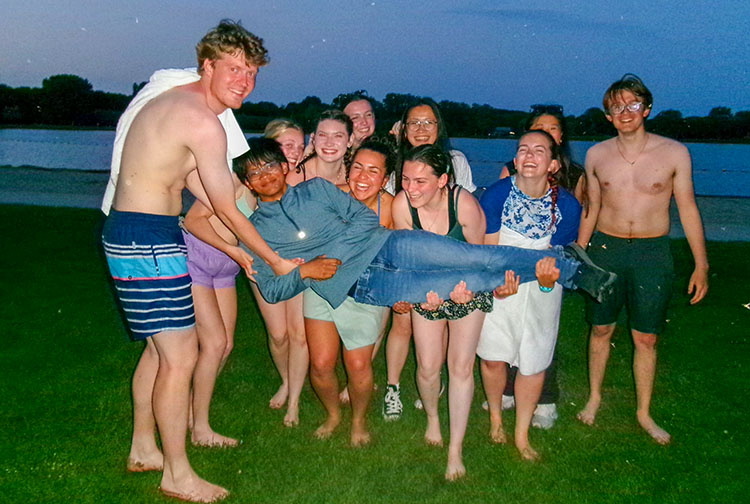 10 young adults in bathing suits and shorts hold another person lying down, on a sandy beach of a lake