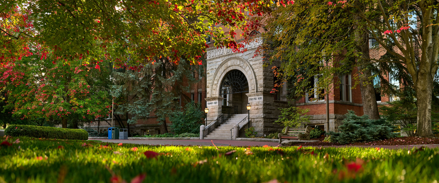 Historic Gonzaga University building framed by vibrant red, orange, and green autumn trees, with a stone archway entrance, campus walkway, bench, and sunlit grass in the foreground.