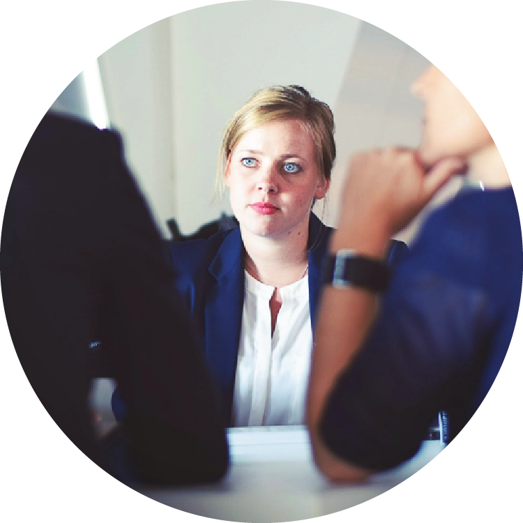 A young woman wearing a blazer sits across from two people, whose backs are to the camera. She appears to listen intently to one of the people across from her. A young woman wearing a blazer sits across from two people, whose backs are to the camera. She appears to listen intently to one of the people across from her.
