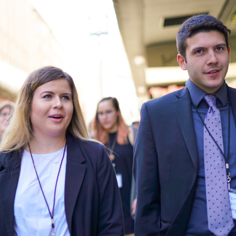 Two students walk side-by-side on the San Francisco trek. The each wear a formal blazer and a lanyard with their name tag on it. Two students walk side-by-side on the San Francisco trek. The each wear a formal blazer and a lanyard with their name tag on it.