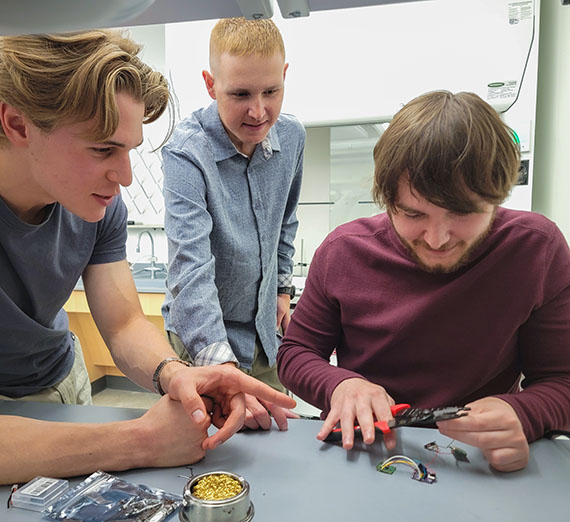 Two students at a table with a professor standing behind, one student with a soldering iron.