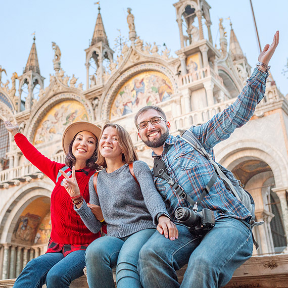 Three smiling young adults pose in front of a historic European cathedral, raising their arms in excitement. 