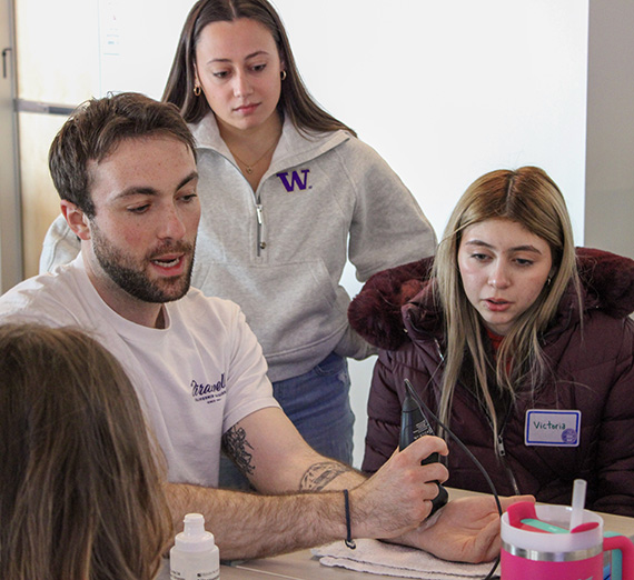A group of five people stand together in a brightly lit hospital hallway, smiling at the camera. Two wear stethoscopes and medical attire, one wears a Gonzaga University sweatshirt, and others are dressed in casual or professional clothing, representing a mix of students and healthcare professionals.
