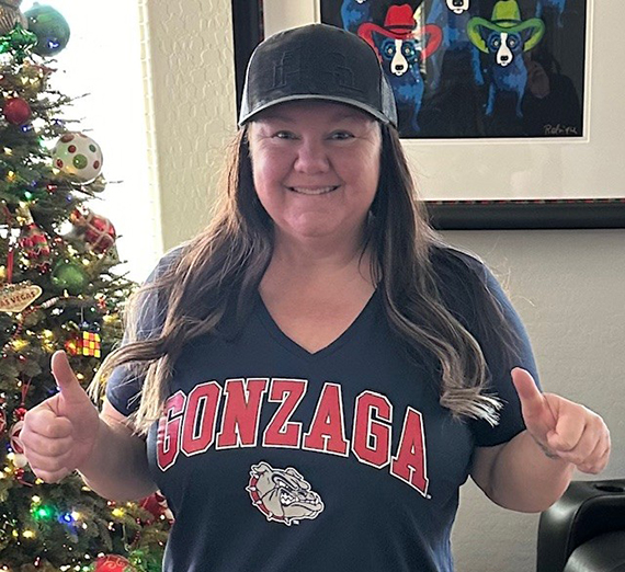 female with brown hair wearing black baseball hat and Gonzaga t-shirt giving a thumbs up with Christmas tree and a painting of 3 dogs in the background