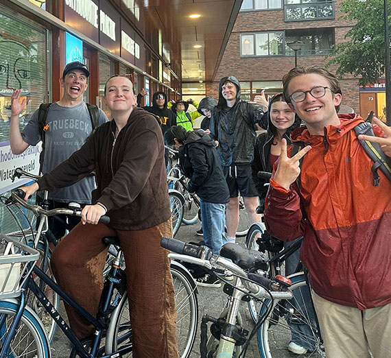 A group of students standing next to bicycles wave to the camera outside a store