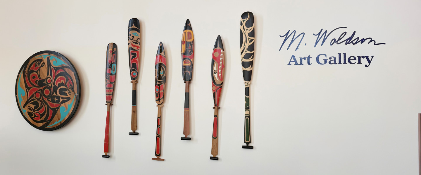 The M. Woldson Gallery logo on a wall with canoe paddles painted with Native American motifs.