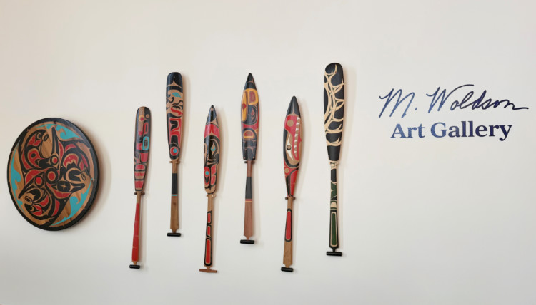 M-Woldson-Gallery-Cover-Image-750x428.jpg The M. Woldson Art Gallery logo with canoe paddles and a shield painted in Native American motifs displayed next to it.