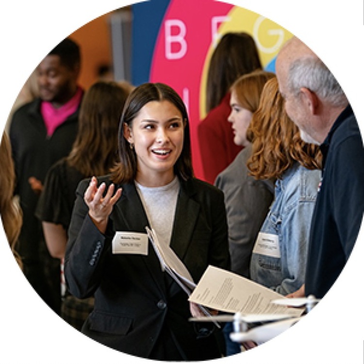 A young woman in professional attire speaks enthusiastically with an older man at a networking event or career fair. She is holding papers and wearing a name tag, surrounded by other attendees in a colorful, busy setting. A young woman in professional attire speaks enthusiastically with an older man at a networking event or career fair. She is holding papers and wearing a name tag, surrounded by other attendees in a colorful, busy setting.
