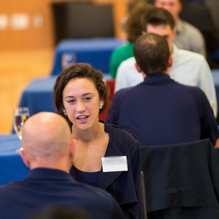 A young woman practices her interview answer with a mock interviewer sitting opposite her at a table. Visible behind her are two more tables in the row with students and mock interviewers similarly interacting. A young woman practices her interview answer with a mock interviewer sitting opposite her at a table. Visible behind her are two more tables in the row with students and mock interviewers similarly interacting.