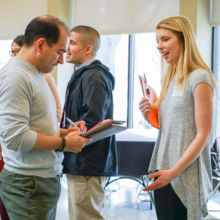 A young woman holds an orange folder as she chats with an employer recruiter at the Education Career Fair. A young woman holds an orange folder as she chats with an employer recruiter at the Education Career Fair.