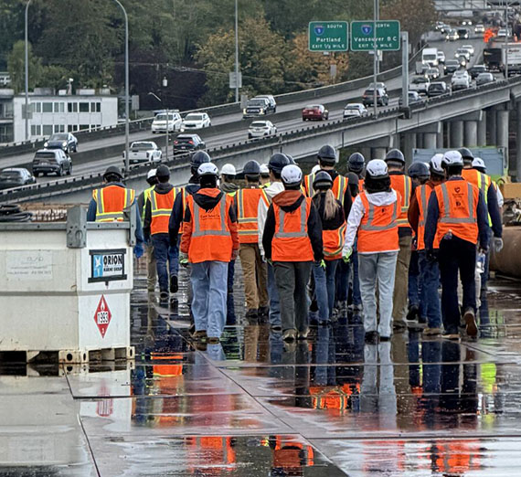 Two dozen people stand together near a freeway wearing safety gear Two dozen people stand together near a freeway wearing safety gear