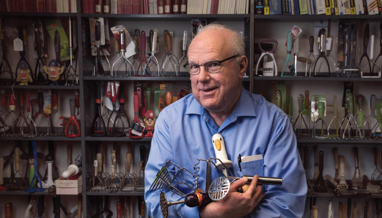 Older man with blue shirt holding potato mashers standing in front of a bookshelf lined with hundreds of potato mashers Older man with blue shirt holding potato mashers standing in front of a bookshelf lined with hundreds of potato mashers