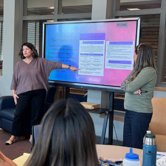 Two women stand in front of a screen presenting to a group of people