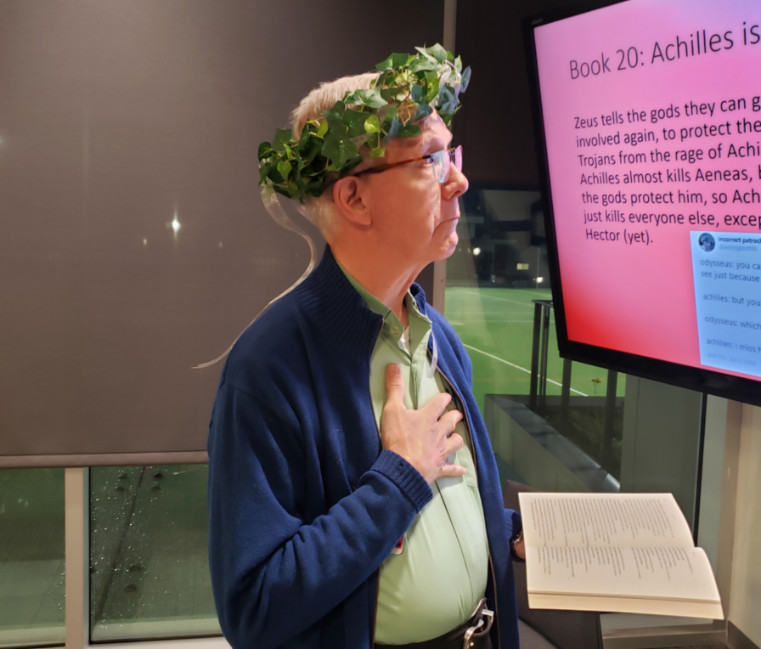 Fr. Ken Krall wearing a laurel wreath on his head holding a book with his other hand over his heart. 