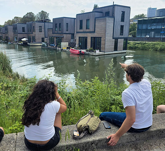 Students sit on the edge of a waterway looking at a row of gray buildings floating on platforms in the middle of the water
