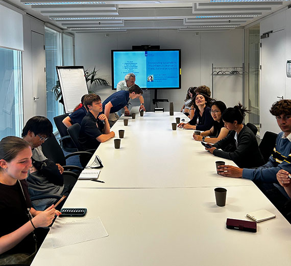 A dozen students are visible at a conference room table preparing for a meeting