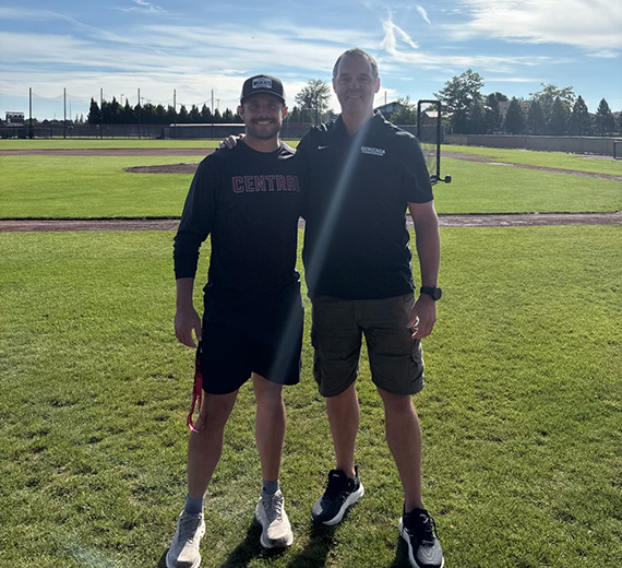 Two men standing on baseball field both wearing shorts and dark tops