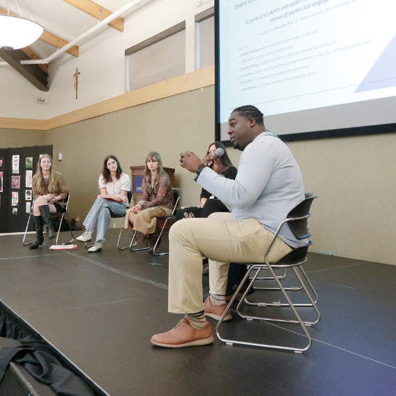 Man sits on stage with microphone with a group of students