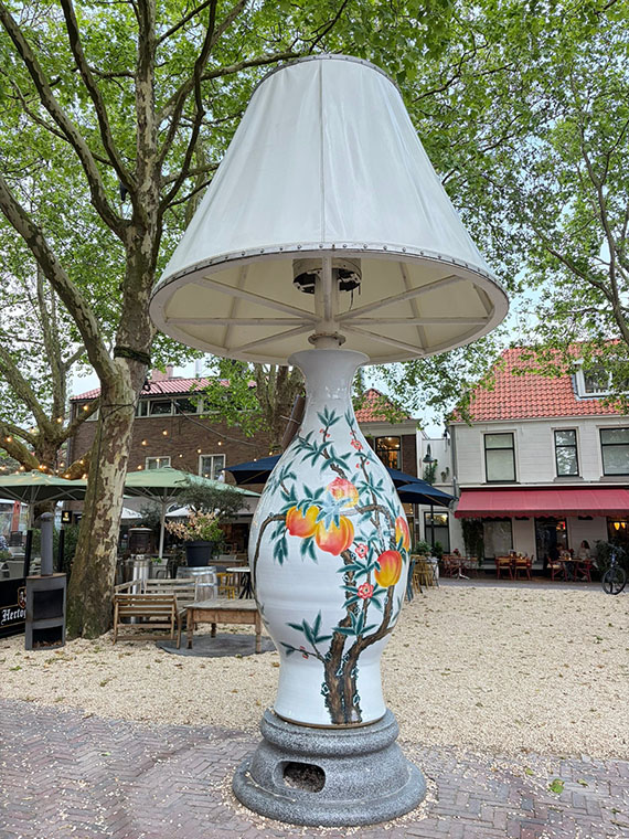 Outdoor sculpture of a table lamp, ceramic base with  Japanese-style illustration of peaches on branches, with a large fabric lamp shade. No people to scale but some outdoor tables and chairs look small behind it.
