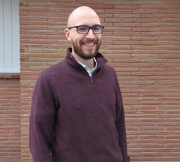 man with glasses wearing a burgundy sweater standing with hands in pocket and brick building in the background