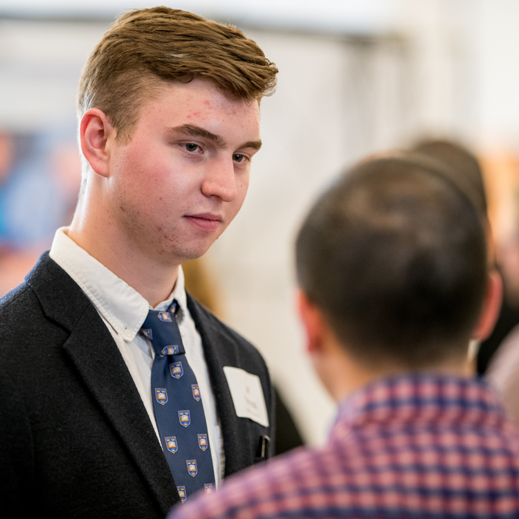 A young man in a formal blazer and tie listens to a recruiter in front of him. The recruiter has his back to camera. A young man in a formal blazer and tie listens to a recruiter in front of him. The recruiter has his back to camera.