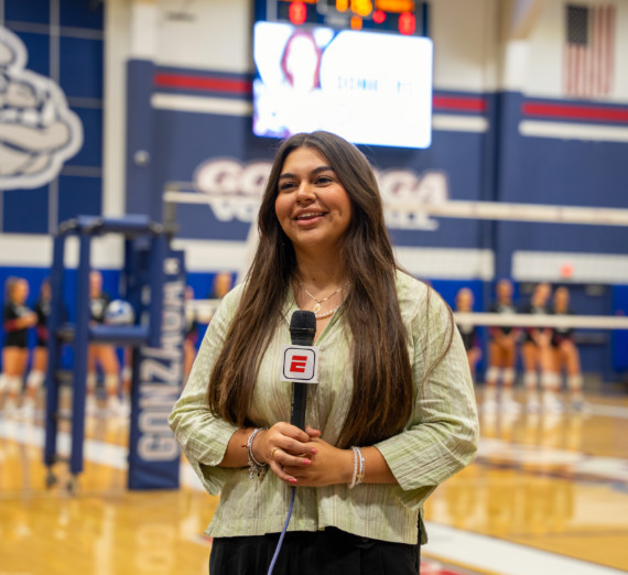 Broadcast journalism student reporting from a Gonzaga University volleyball game, holding an ESPN microphone on the court with players and scoreboard visible in the background.