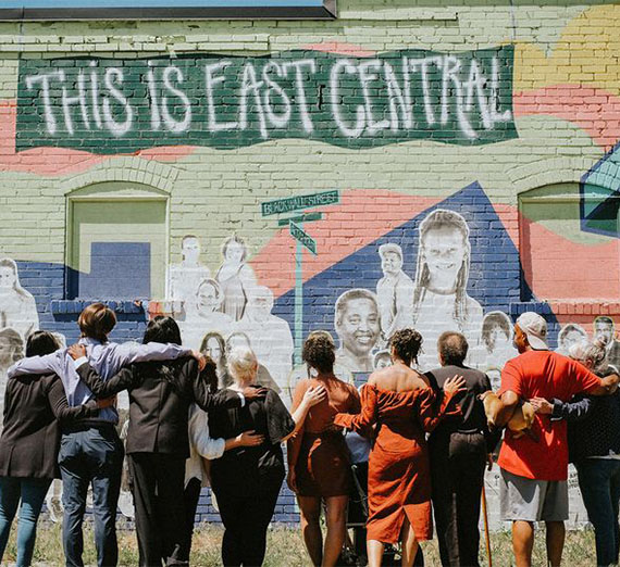 A group of people with their arms around one another standing in front of a mural that displays community members and 