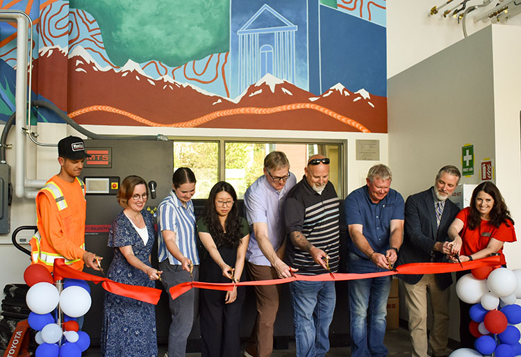 Nine adults stand behind a red ribbon with large scissors in hand.