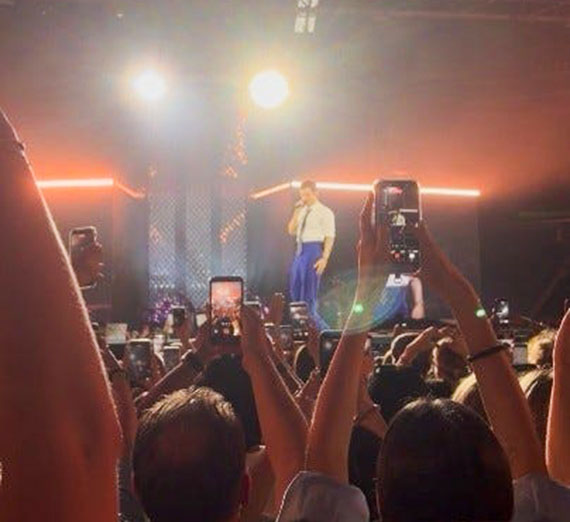 A singer stands on a stage in the distance while fans hold their phones up to catch a photo or video