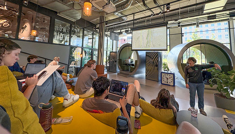 Students sit on large yellow steps in a communal classroom and study space watching a teacher