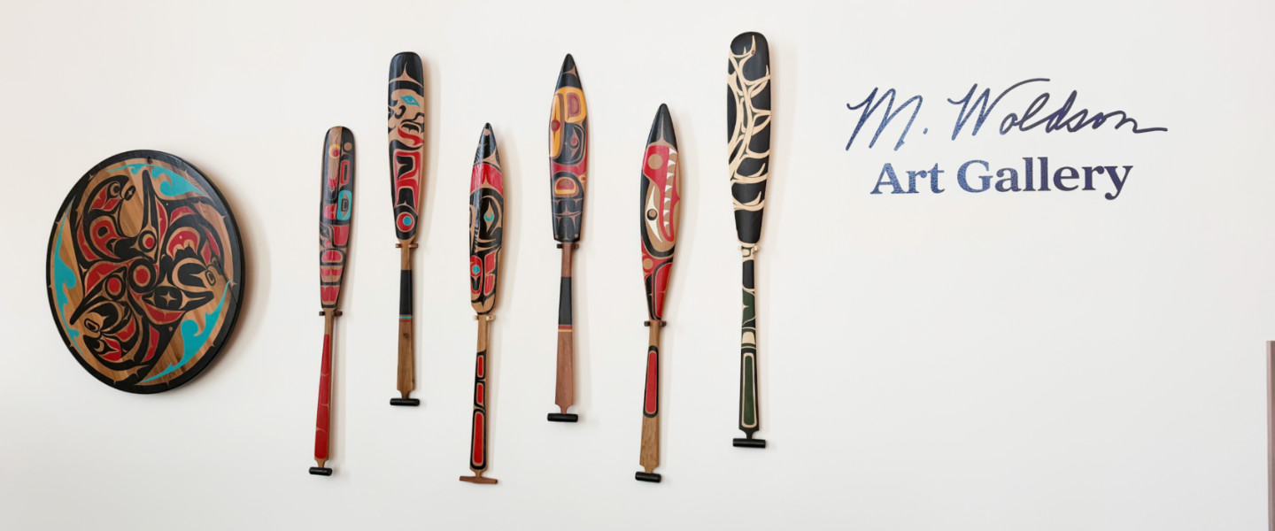 The M. Woldson Gallery logo and a row of Native American canoe paddles. 
