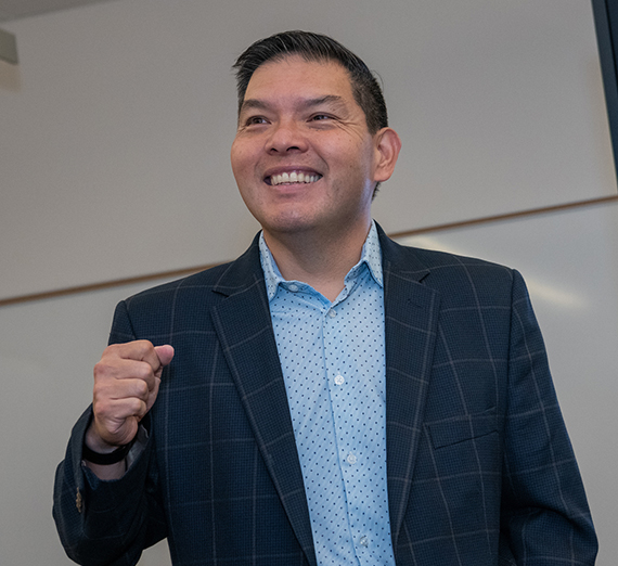 A man with arms outstretched as he speaks at head of a classroom