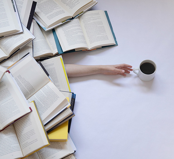 Person under a pile of books reaches for a cup of coffee.