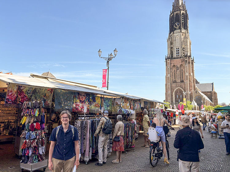 A student stands in the left of the photo smiling at the camera while behind him people look at stalls in an open-air market