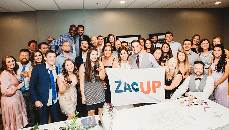 A group of wedding guests indoors, smiling and holding a 'Zag Up' flag. A group of wedding guests indoors, smiling and holding a 'Zag Up' flag.