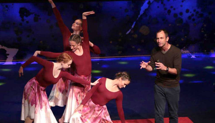 Four dancers in red long sleeved shirts and pink/white skirts enact dance that resembles the movements of animals. Professor Brook Swanson speaks to a crowd in front of them. Four dancers in red long sleeved shirts and pink/white skirts enact dance that resembles the movements of animals. Professor Brook Swanson speaks to a crowd in front of them.