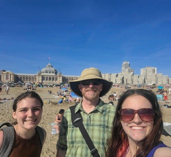 Three students in sunglasses and sun hats stand on a beach with a large ornate boardwalk building behind them.