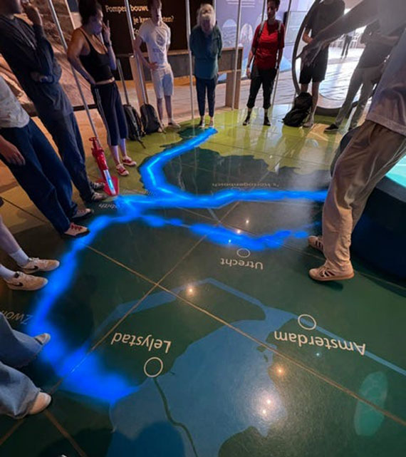 Many students stand around a mosaic on the floor creating a map of the region with a stripe representing water in blue light