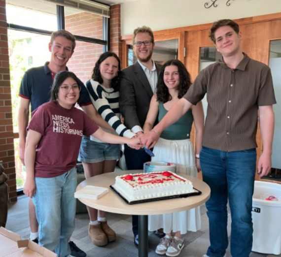 Group of Gonzaga University History Department students gathered around a table cutting a celebratory cake during a Phi Alpha Theta event. One student wears a maroon T-shirt with the text ‘America Needs Historians.’ Group of Gonzaga University History Department students gathered around a table cutting a celebratory cake during a Phi Alpha Theta event. One student wears a maroon T-shirt with the text ‘America Needs Historians.’
