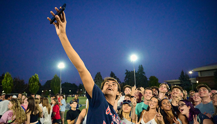A man holds up a cell phone to take a selfie in front of a group of students outside under a dark sky A man holds up a cell phone to take a selfie in front of a group of students outside under a dark sky