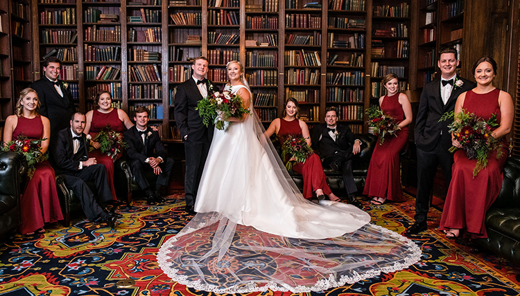 A bride and groom standing in a library, surrounded by books and in the company of bridesmaids - in red dresses - and groomsmen - in black suits. A bride and groom standing in a library, surrounded by books and in the company of bridesmaids - in red dresses - and groomsmen - in black suits.