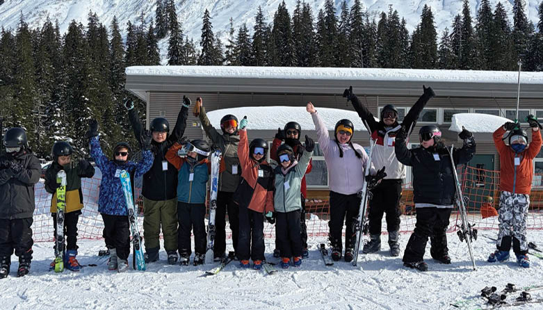 A group of Indigenous youth skiers pose in front of treed, snowy mountain A group of Indigenous youth skiers pose in front of treed, snowy mountain
