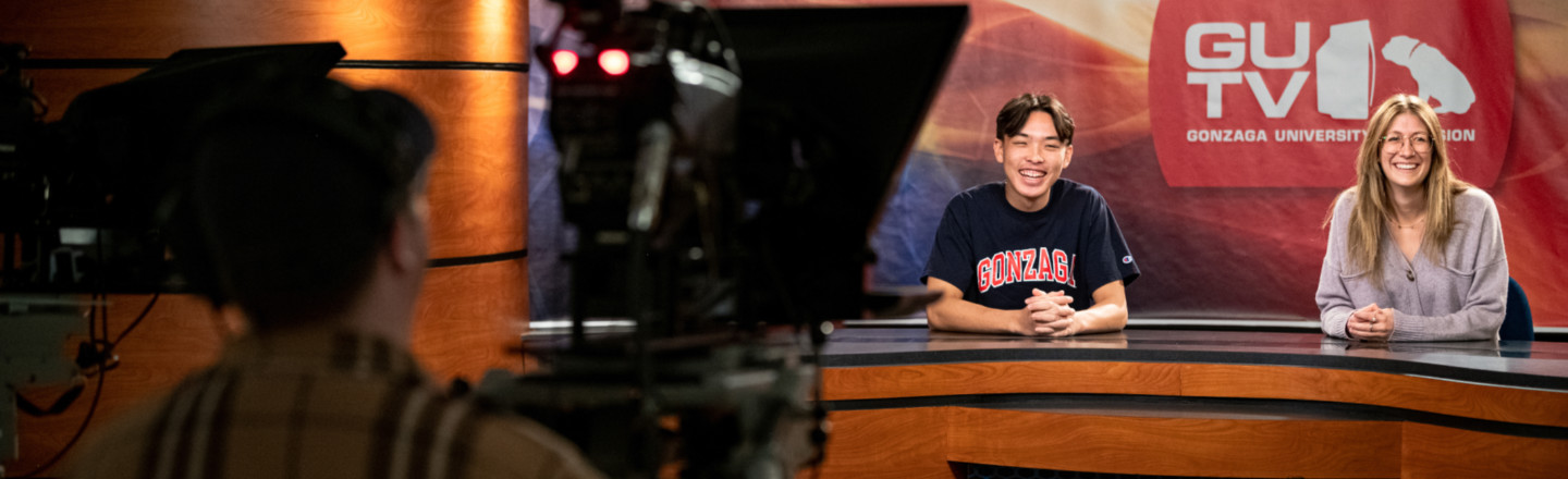 A TV studio with an out of focus camera person in the left foreground and two students in the right background at a broadcasting desk with a sign behind them reading 