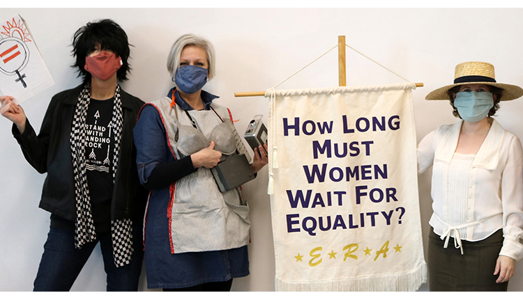 three women in costumes and a banner that reads How Long Must Women Wait for Equality three women in costumes and a banner that reads How Long Must Women Wait for Equality