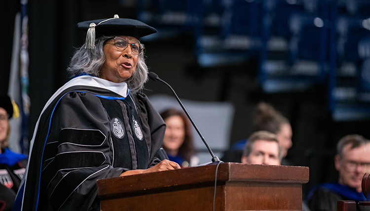 A woman in graduation regalia speaks at a podium A woman in graduation regalia speaks at a podium