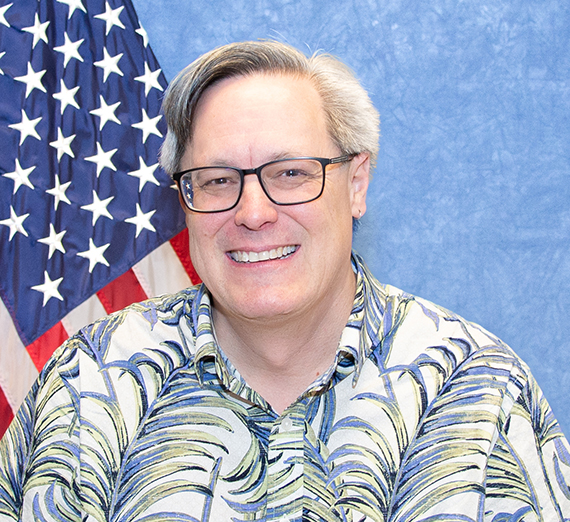 Brian Heckard wearing glasses, a button up with a leaf pattern, and in front of a blue background with an American flag in the corner. 