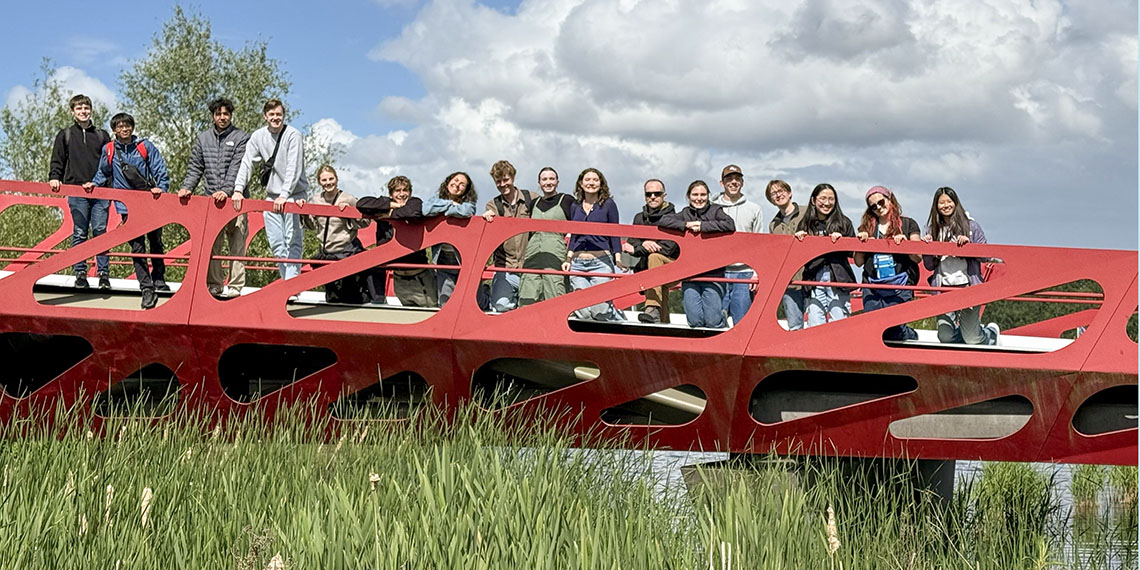 18 students on a red metal bridge over grass